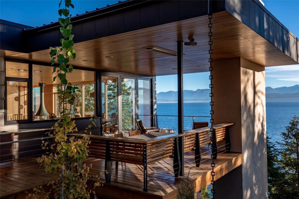 Porch with views of a Montana lake, and through floor-to-ceiling windows into a living room with modern fireplace.