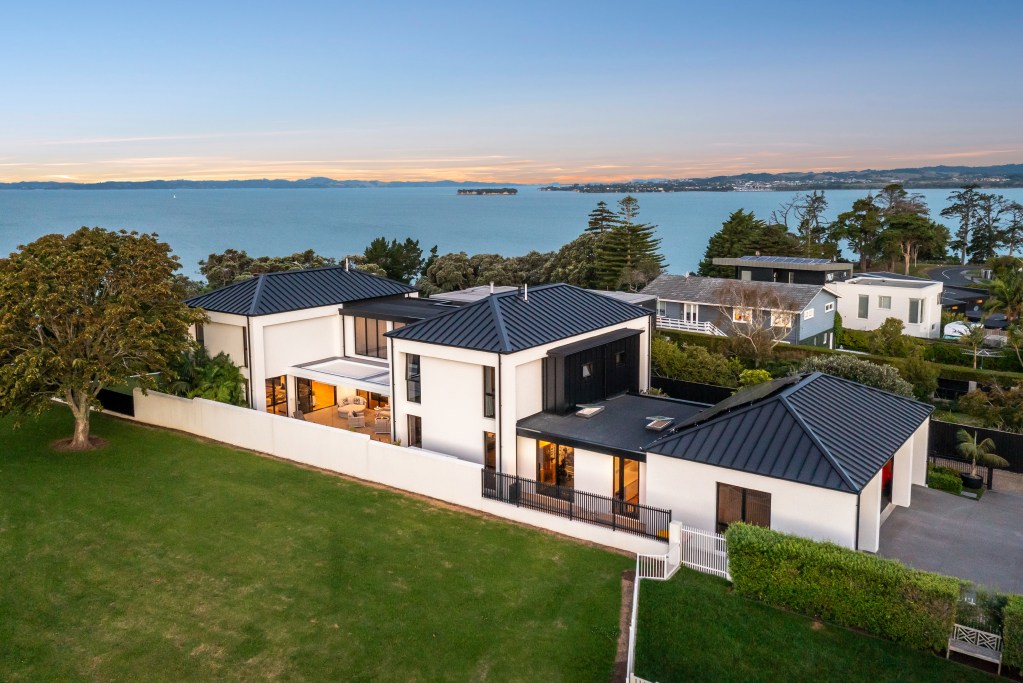 Aerial view of a contemporary waterfront residence featuring multiple black metal rooflines, a large lawn, and panoramic bay views at sunset.
