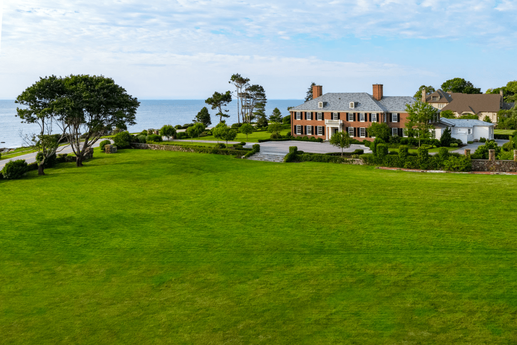 Aerial view of a stately brick residence overlooking the coast, featuring a wide manicured lawn, formal landscaping, and unobstructed water views.