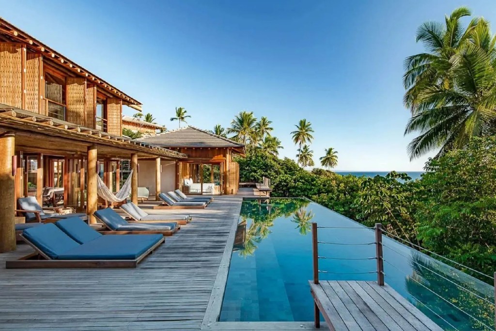 Tropical luxury villa featuring an infinity pool, expansive wooden deck, lounge chairs, and lush palm-filled gardens with ocean views in the distance. Itacare, Brazil.
