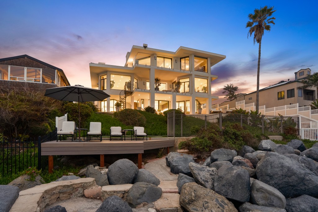 Contemporary oceanfront residence with floor-to-ceiling windows, multiple terraces, and coastal views, photographed from the shoreline at sunset.
