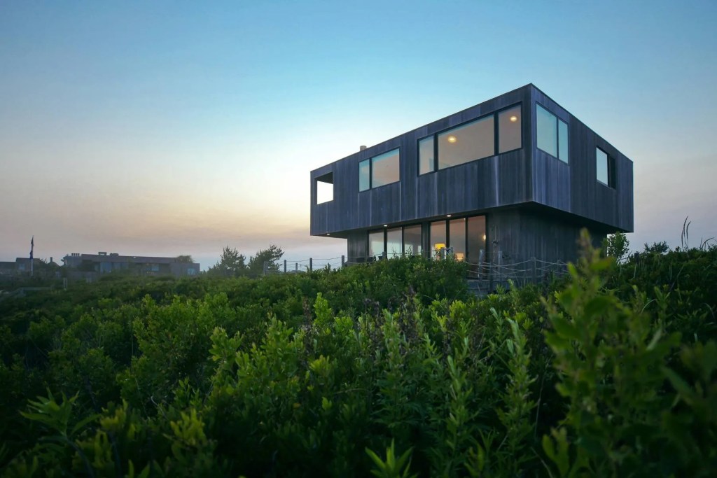 Modern coastal home with a dark wood exterior and expansive windows, surrounded by natural dunes and lush greenery at sunset. Sagaponack, New York.