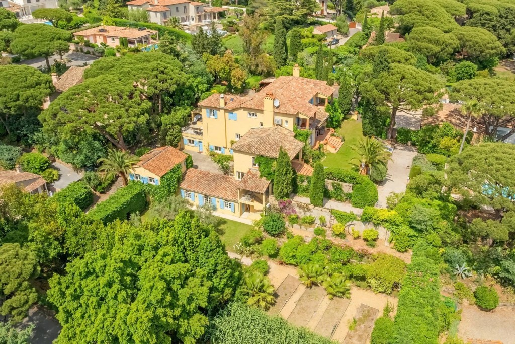 Aerial view of a sprawling Mediterranean estate with terracotta roofs, lush landscaped gardens, and tall umbrella pine trees throughout the surrounding neighborhood. Sainte Maxime, France.