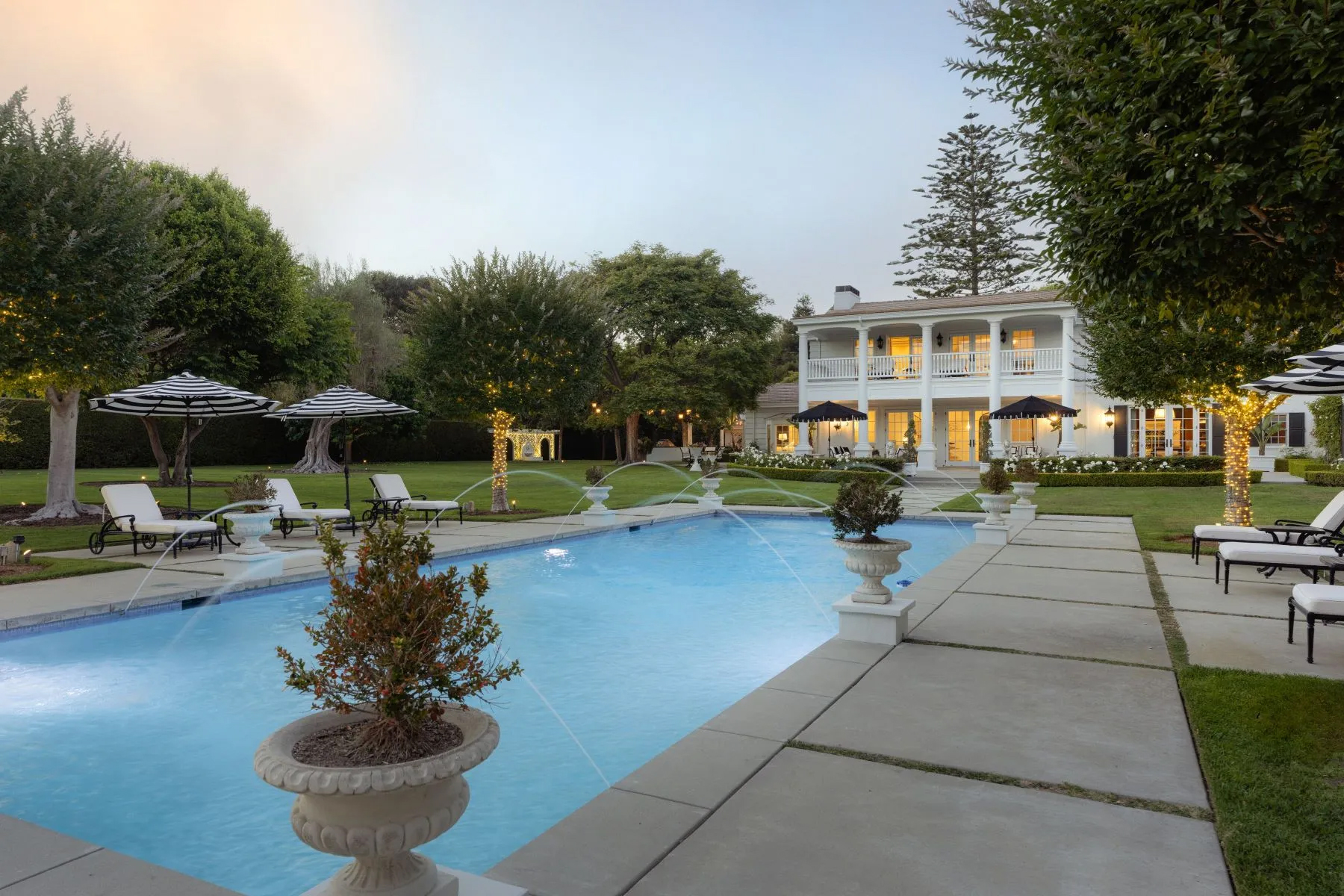 Elegant white estate overlooking a large rectangular swimming pool with decorative water fountains, lounge chairs, and manicured lawns illuminated at dusk.