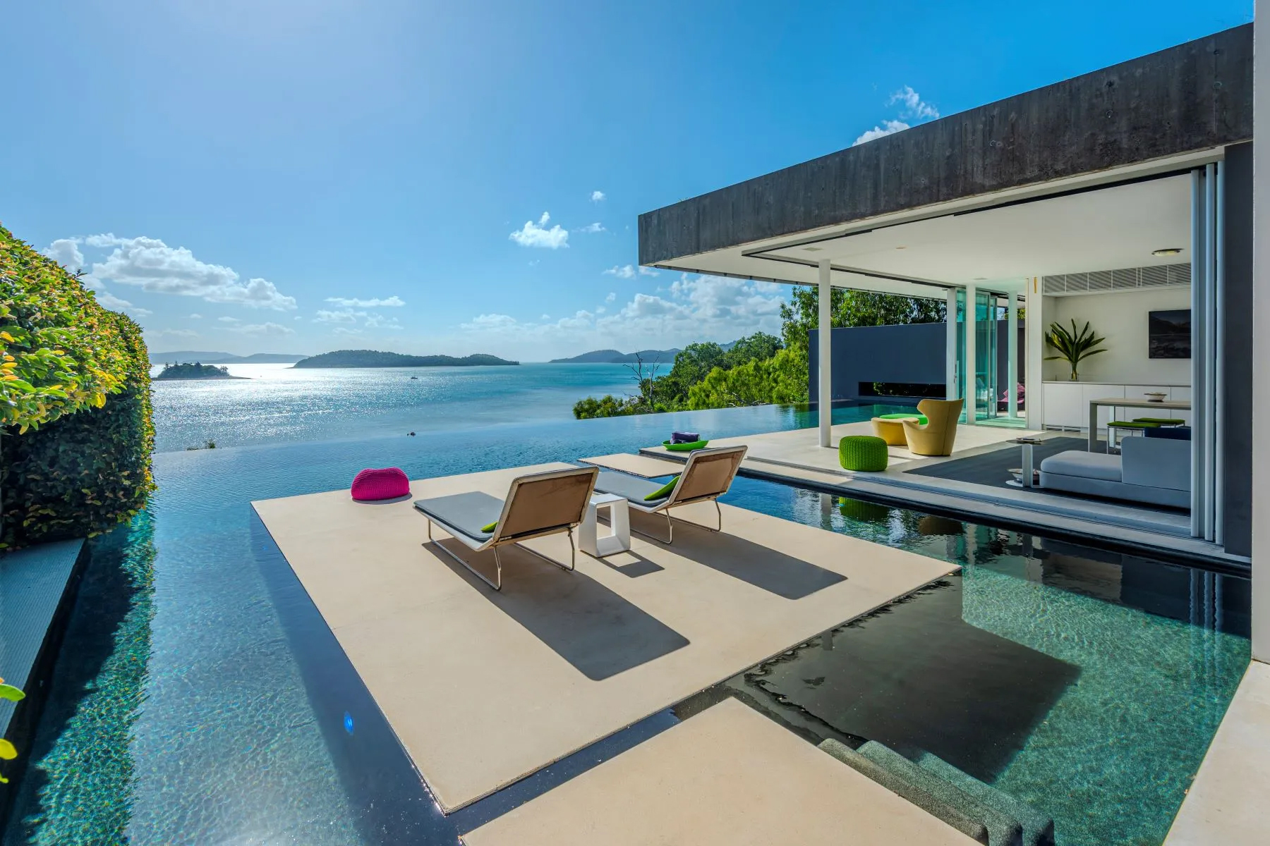 Modern waterfront villa with an infinity pool, outdoor lounge area, and panoramic views of a blue ocean dotted with small islands.