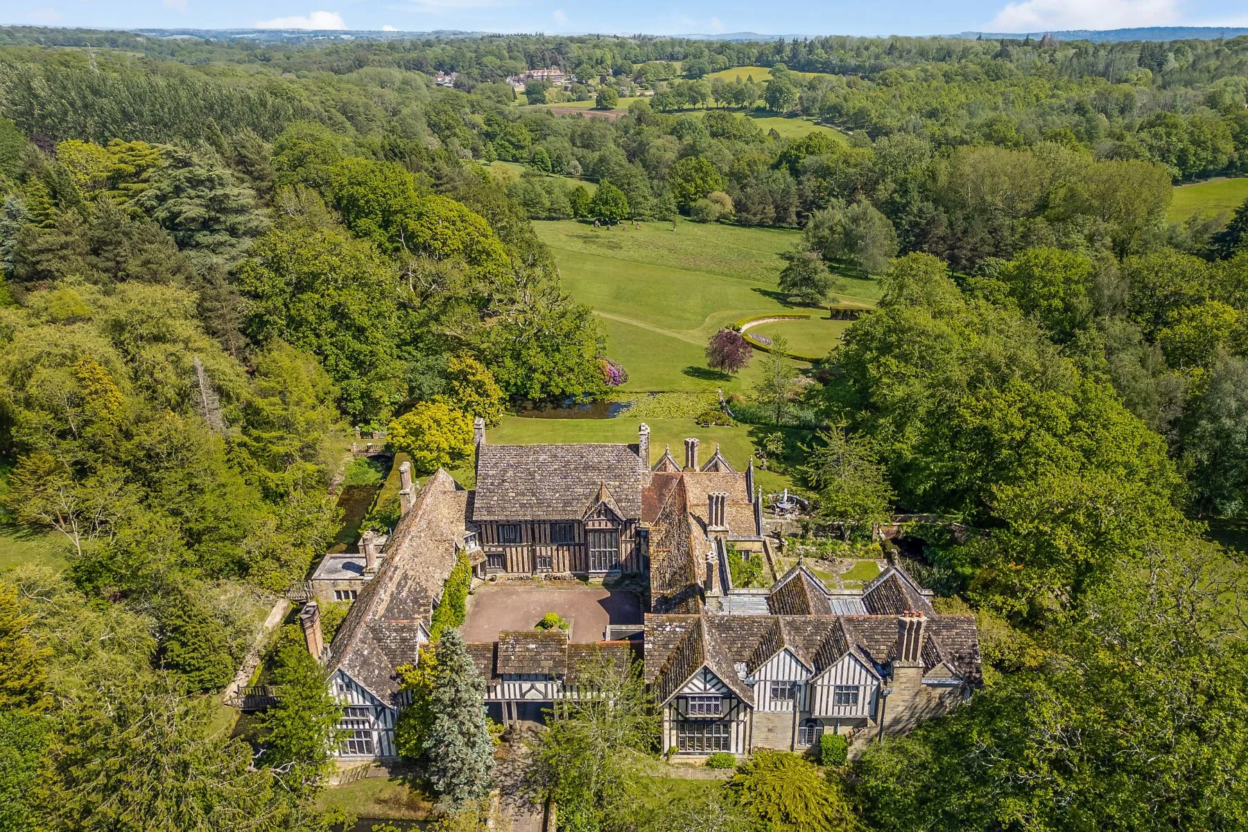 Aerial view of a historic countryside estate surrounded by mature trees, expansive lawns, and rolling green landscape extending into the distance.