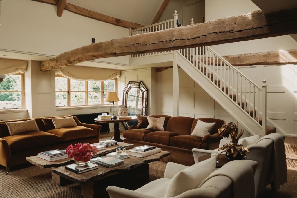 reverse perspective of sitting room in Cotswold's home, with rough exposed wood beams, plush velvet seating, side table with lighting, and standing mirror in the background with stairs leading to another floor
