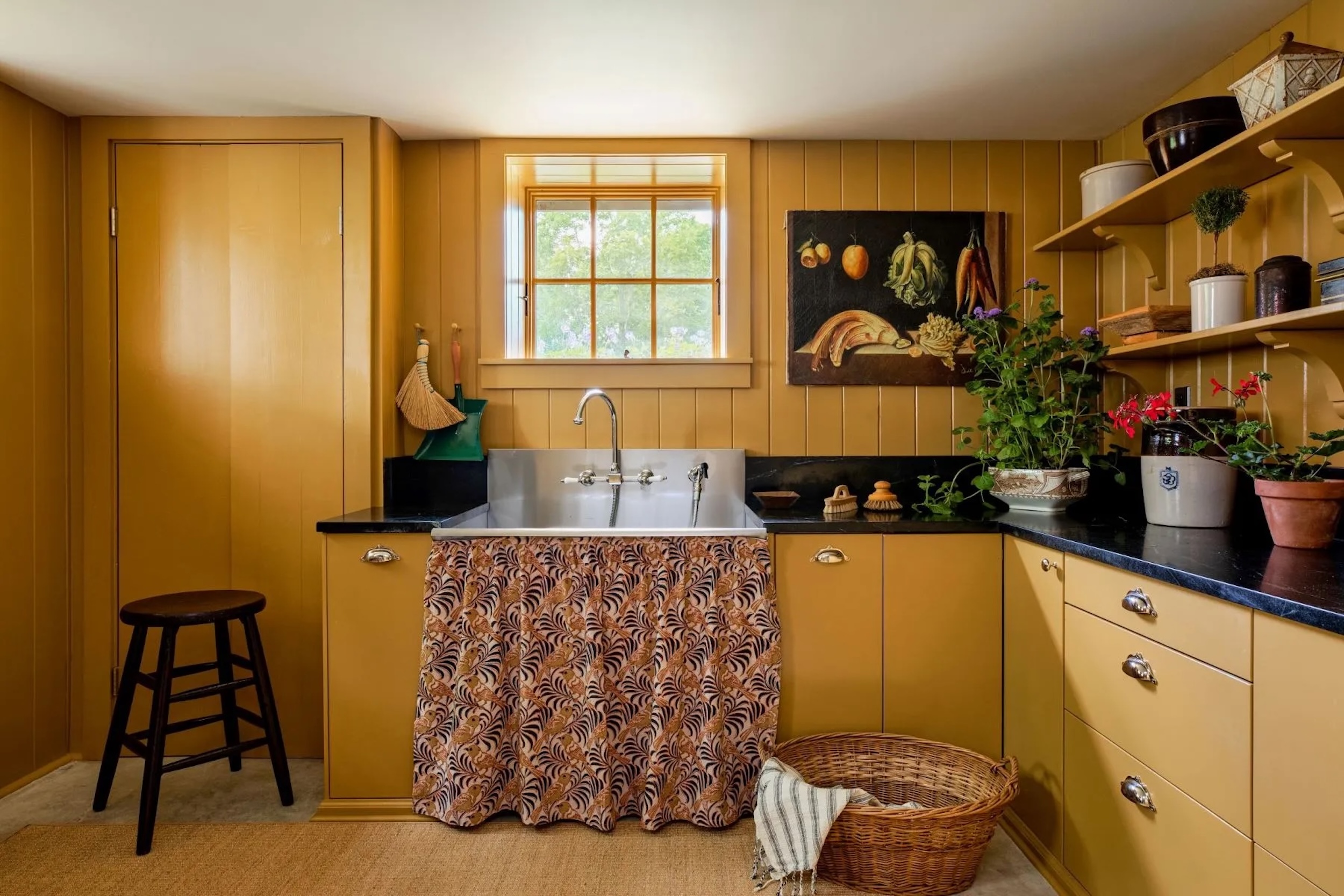 rustic yellow kitchen in Hudson Valley, NY home, featuring farm sink with matching fabric skirt, bright cabinets and drawers, matching wood paneled walls, still life art piece, basket on floor, and dark marble countertops with flower pots