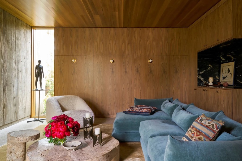 cozy corner in Venice Beach, California home with blue velvet couch, plush white chair, large stone coffee table, plush rug, wood paneling on walls and ceiling, and tall window in the background