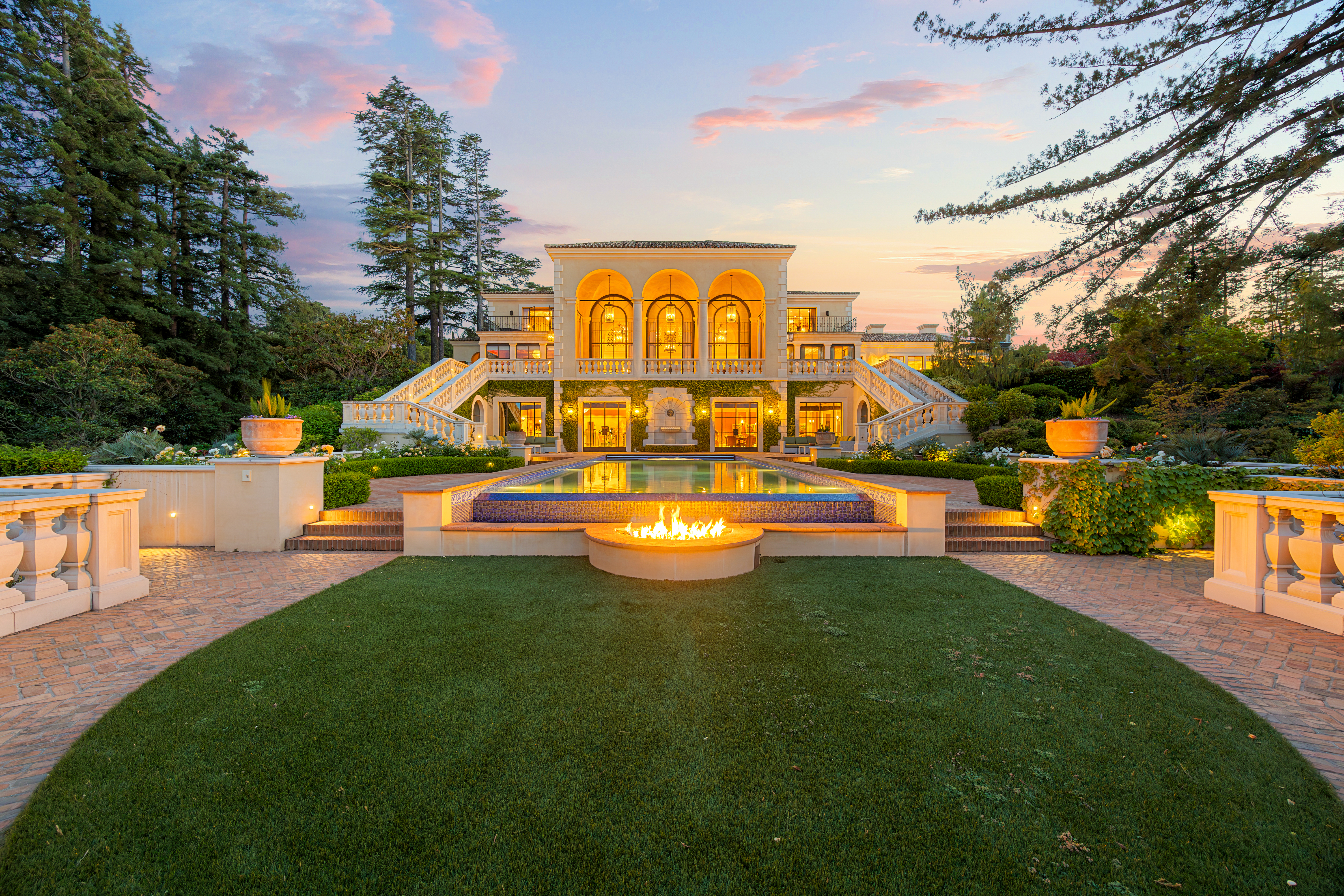 Luxury Mediterranean-style residence at sunset with symmetrical architecture, illuminated interiors, formal gardens, a reflecting pool, and an outdoor fire feature.