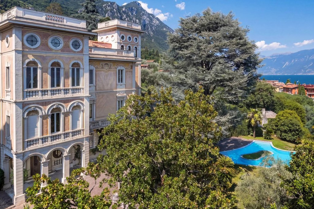 Historic villa overlooking a lake and mountains, featuring ornate architecture, lush gardens, and a curving outdoor swimming pool.