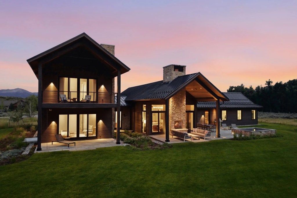 Contemporary two-story home with wood and stone exterior, covered outdoor living area, and landscaped lawn at dusk beneath a pink and blue sky.