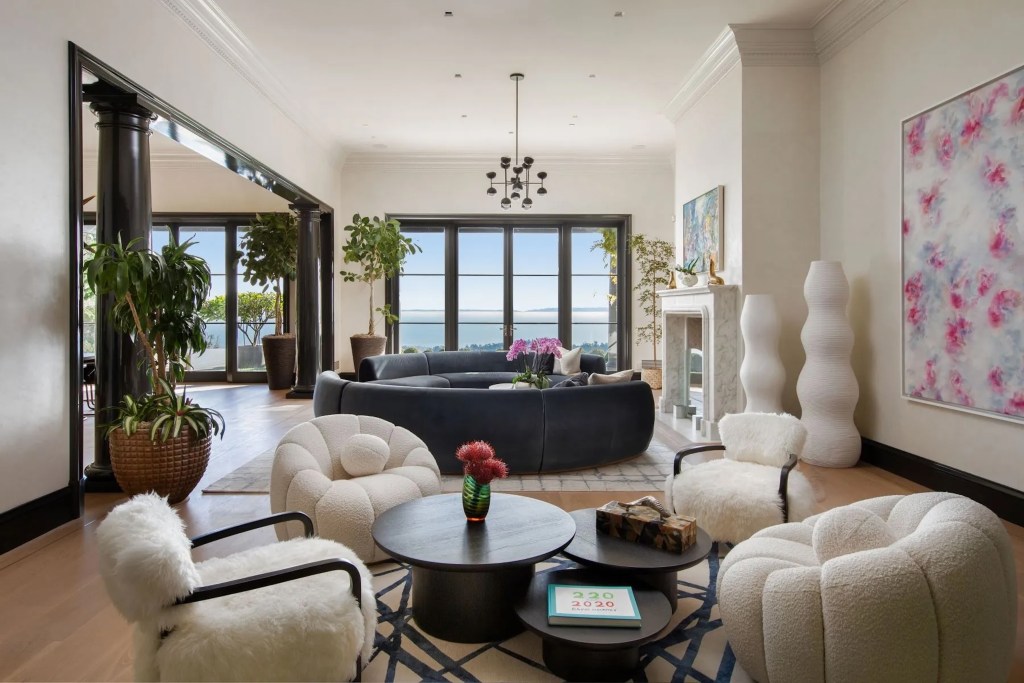 salon in Montecito home with circular couch, modern plush seating, marble fireplace, free standing sculptures, art on walls, and house plants, facing a window that overlooks the ocean