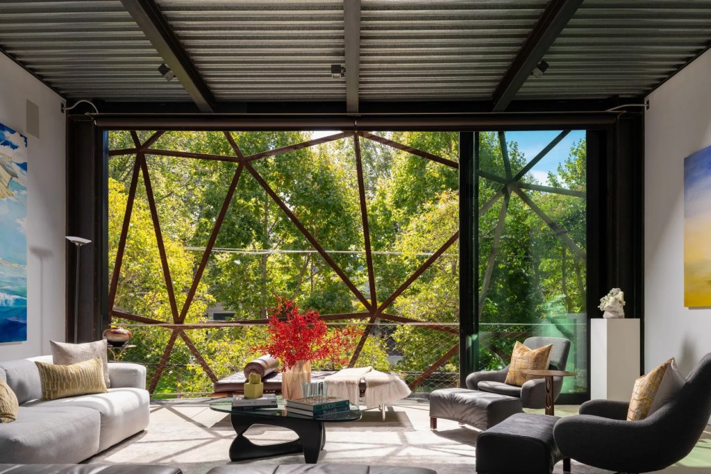 Contemporary living room with seating area and large floor-to-ceiling windows overlooking trees through a geometric metal screen.