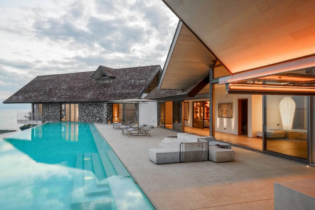 Modern waterfront residence with floor-to-ceiling glass walls, an infinity-edge swimming pool, and expansive outdoor living spaces overlooking the water.