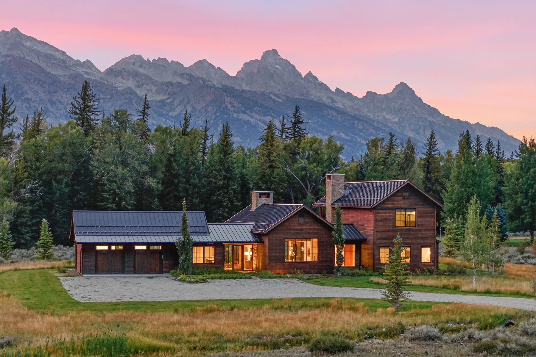 Luxury mountain home surrounded by evergreens and open meadow, with the Teton mountain range rising behind it at sunset beneath a pink sky.