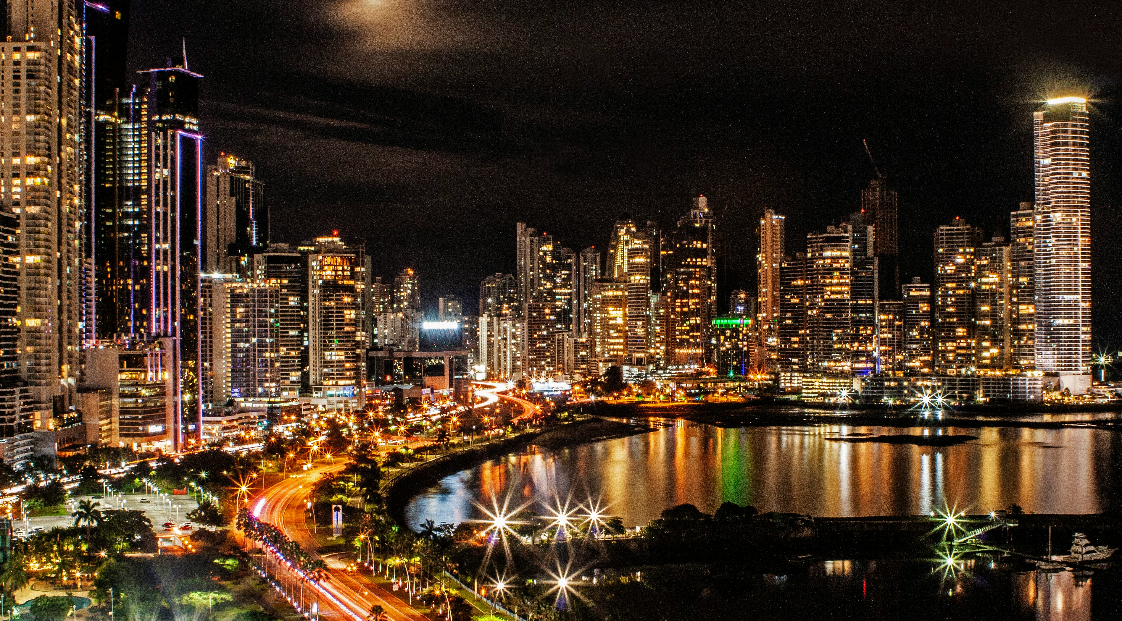 Nighttime view of a modern waterfront skyline with illuminated high-rise buildings reflected in a bay. Curving roads create streaks of light from moving traffic, while city lights and skyscrapers glow against a dark sky.