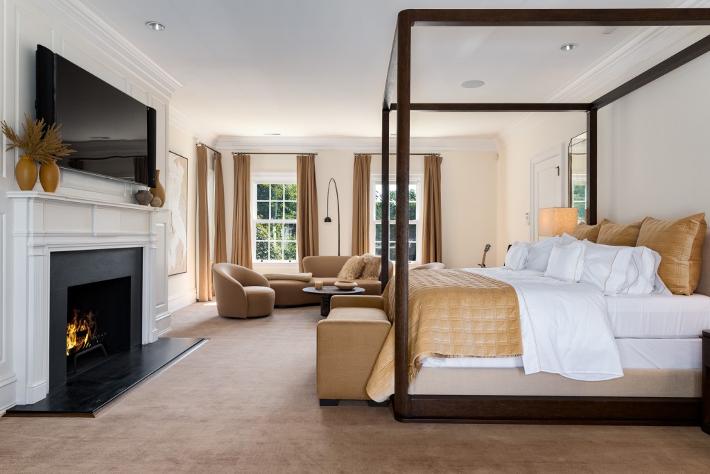 primary suite in Greenwich home with espresso wood framed bed with gold and white bed linens, a fireplace and small seating area with abstract curved sofas and chair