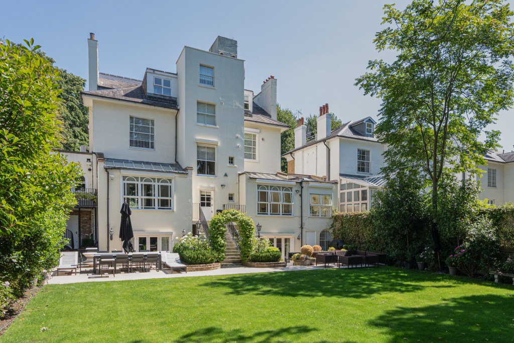 Rear exterior view of a tall white residential property featuring large windows, terraces, outdoor seating areas, and a landscaped garden with mature trees and a spacious green lawn.