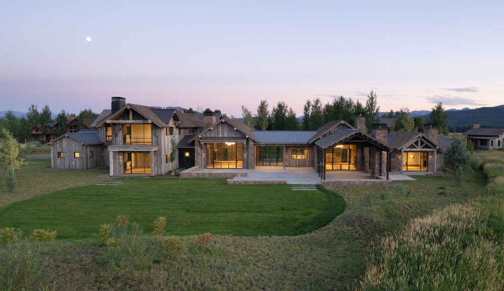 Large wood-and-stone luxury home with expansive windows and covered outdoor living areas, set on a manicured lawn at dusk beneath a pale evening sky.
