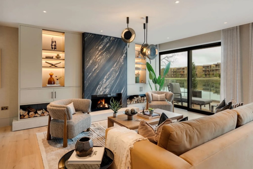 sitting room in Cotswolds, England, with plush seating, black marble fireplace, built-in shelving with recessed lighting, and tree minimalist pendant lights near a window facing a balcony over water