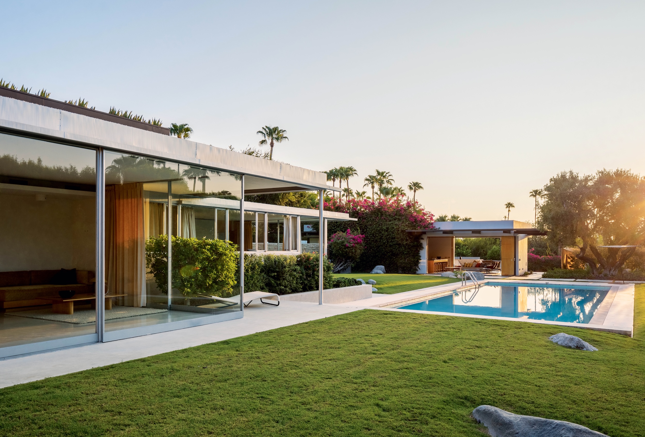 Exterior of The Kaufmann Desert House in Palm Springs, designed by Richard Neutra in 1946, featuring expansive glass walls, modular architecture, and a lush lawn leading to pool with palm trees framing the home