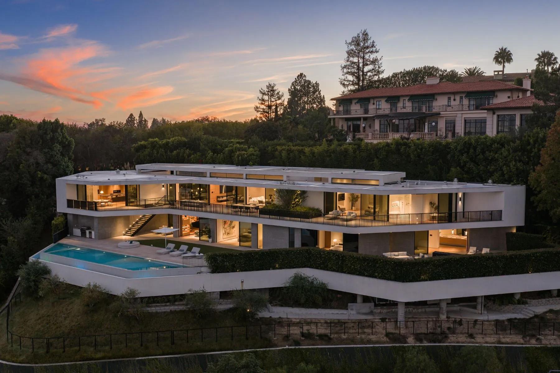 Modern luxury home with floor-to-ceiling windows, outdoor living spaces, and a lighted infinity-edge pool at dusk.