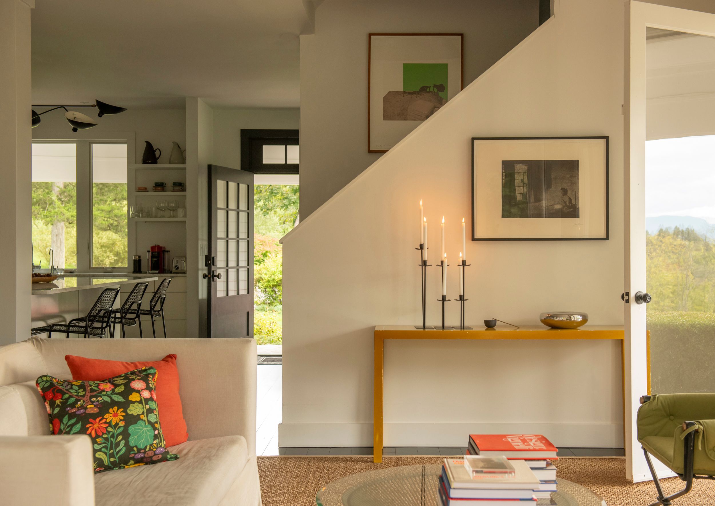 sitting room in Germantown, NY home, with a wall leading up to a staircase and a console table against it with a 6 taper candles, with art prints on walls, and a kitchen counter and open front door in the background