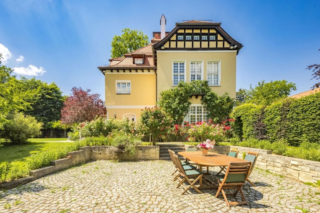 stone patio of Munich home with dining table and chairs, with flourishing landscaping, and yellow Art Nouveau home in the background