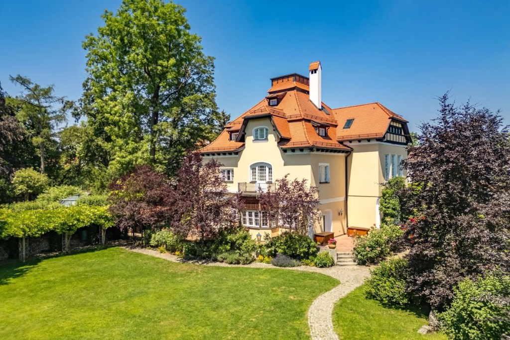 exterior of yellow Art Nouveau villa in Munich, with large trees and verdant landscaping and orange sloped roof