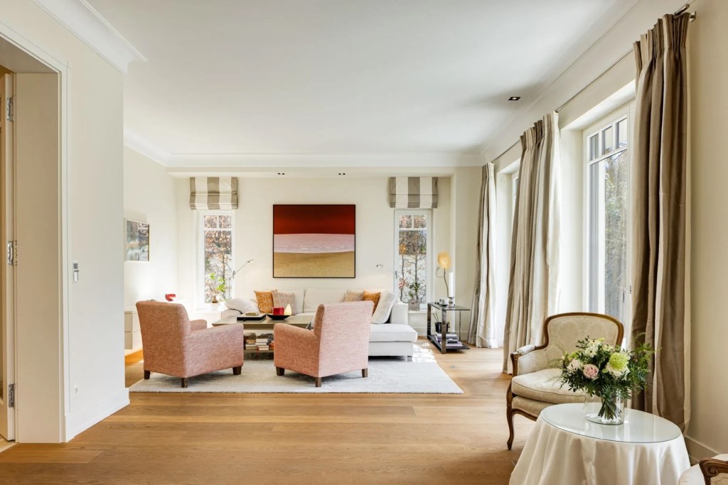 interior of Munich's Villa Elysee, with white walls, large windows, and a seating area with white sofa and two pink chairs, with art in the background depicting a similar color scheme