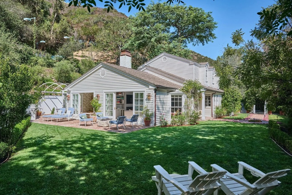 Rear exterior of a white shingle-style home with a brick patio, outdoor lounge seating, manicured lawn, mature trees, and hillside landscaping surrounding the property.